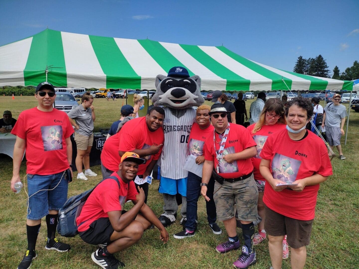 Group of people in red shirts posing with a mascot in front of a green and white tent.