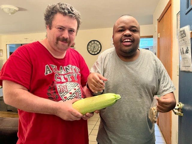 Two men smiling, holding a large zucchini in a kitchen.