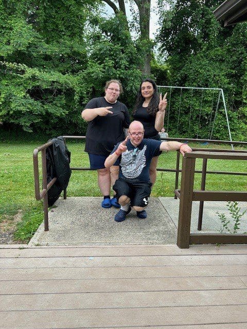 Three people posing happily on a small wooden bridge in a green outdoor setting.