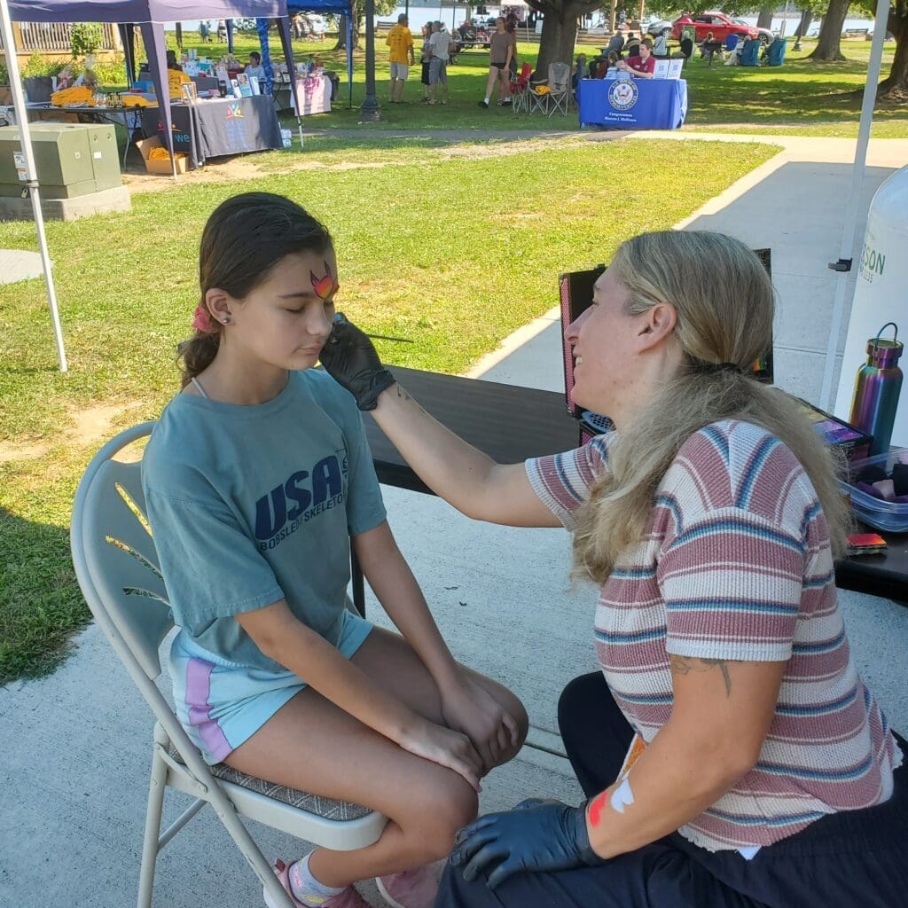 A woman applying face paint to a young girl at an outdoor event.
