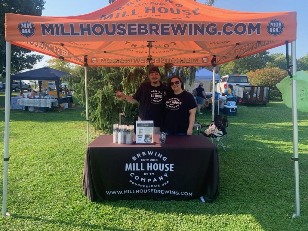 Two people behind a Mill House Brewing booth at an outdoor event.