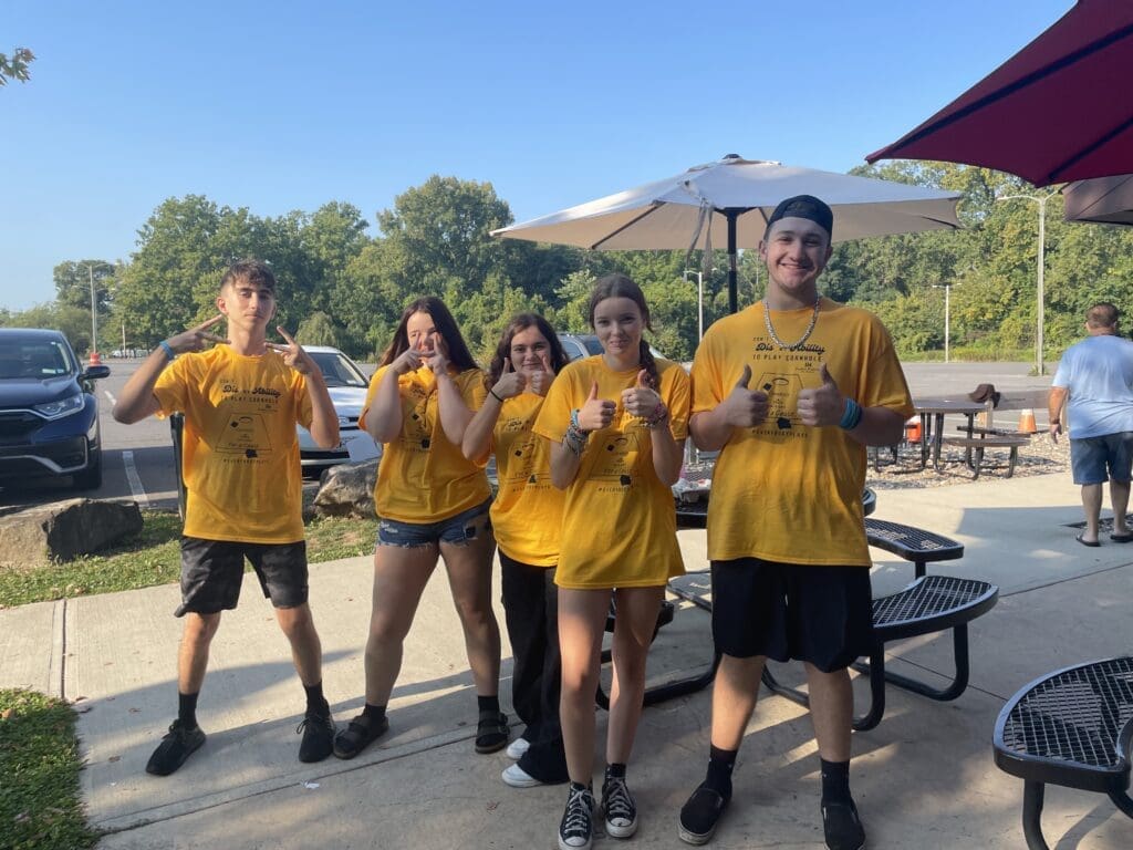 Five people in yellow shirts posing outdoors near picnic tables and umbrellas.