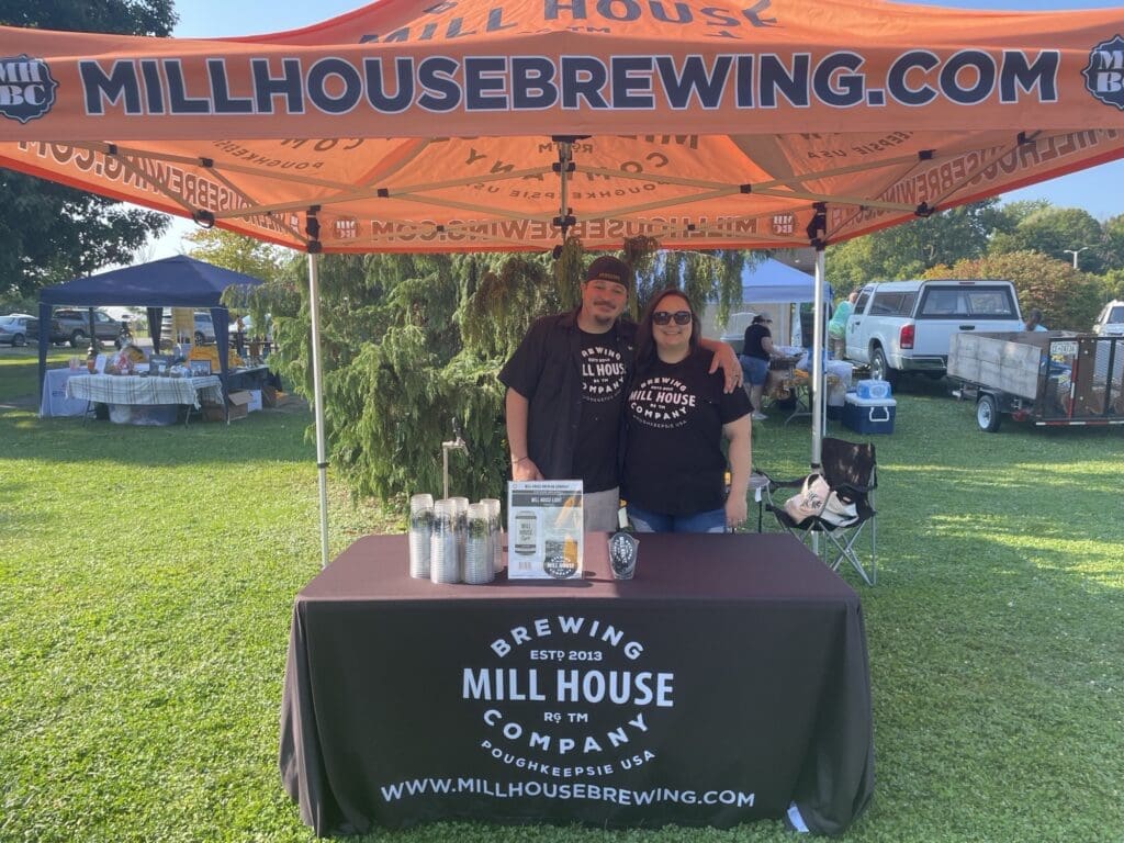 Two men at a Mill House Brewing Co. booth under an orange tent.