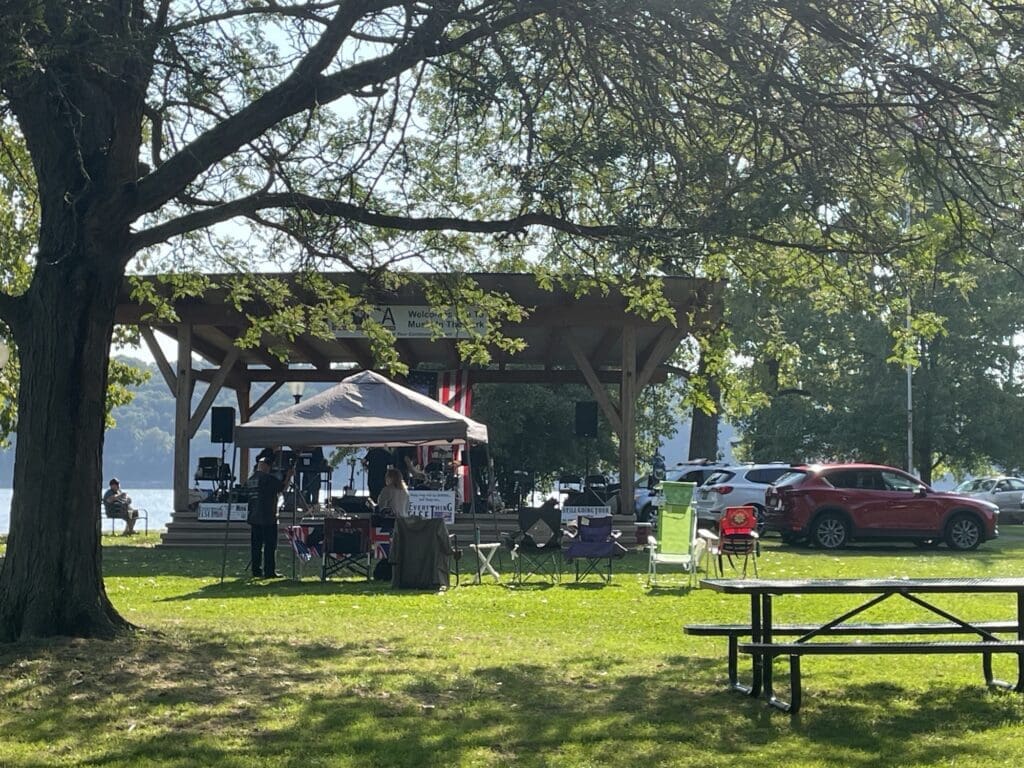 People gathered outdoors under trees with a tent and picnic tables nearby.
