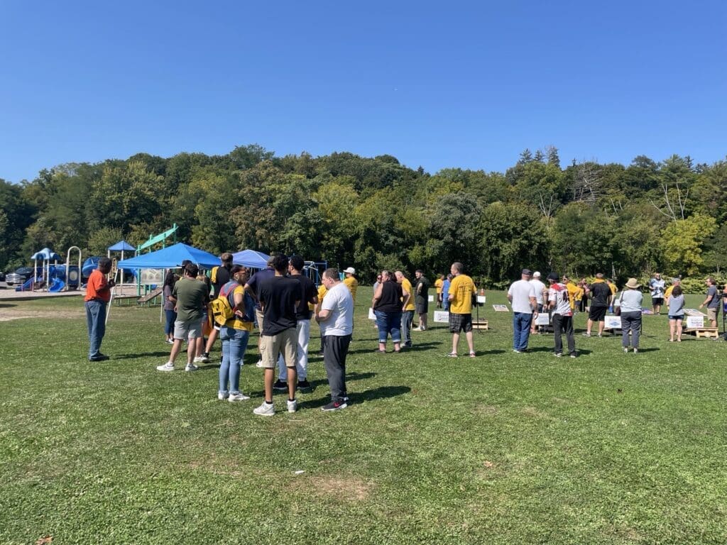 People gathered outdoors on a sunny day with tents and trees in the background.