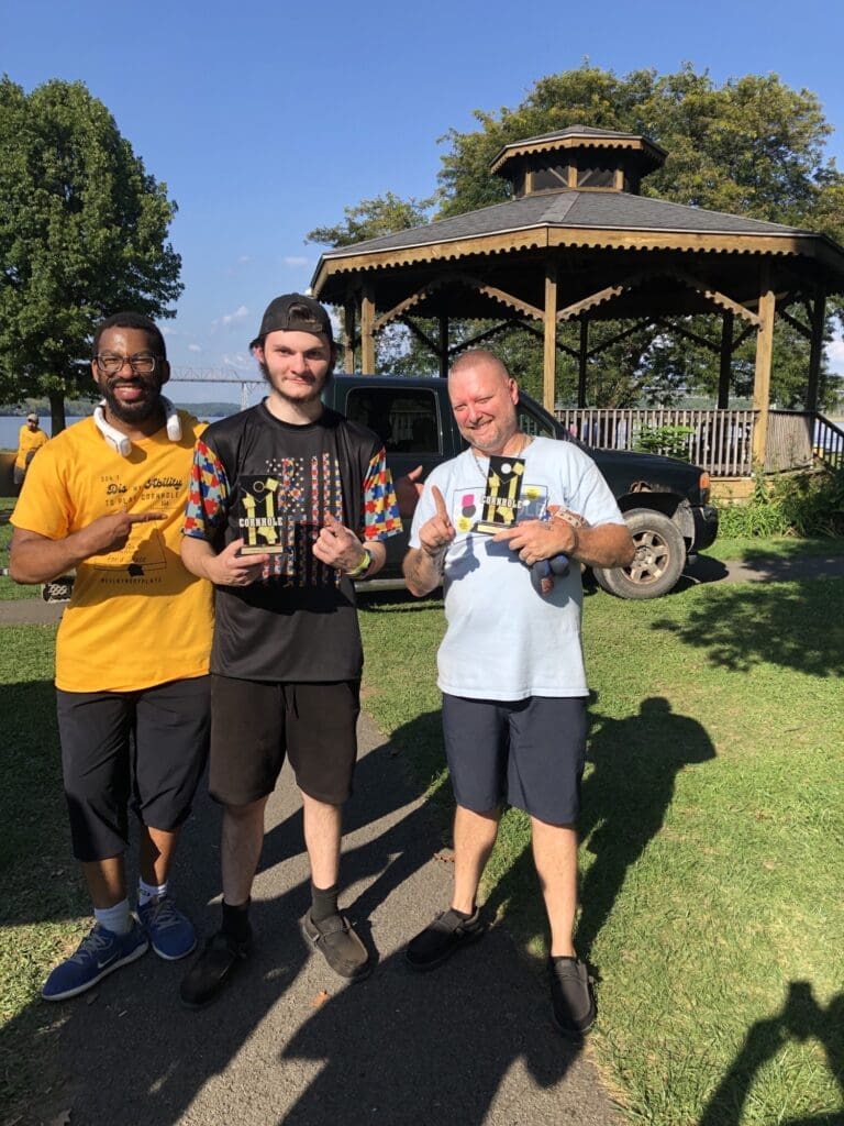 Three men smiling and holding trophies outdoors near a gazebo.