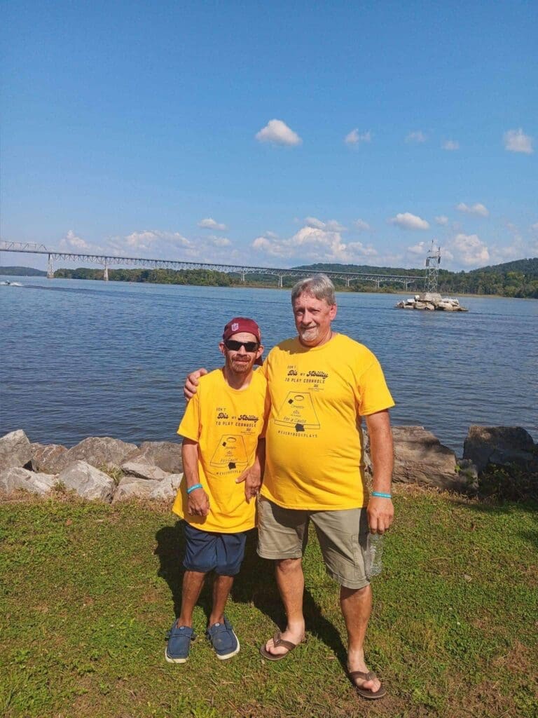Two men in matching yellow shirts pose by a lakeside with a bridge in the background.