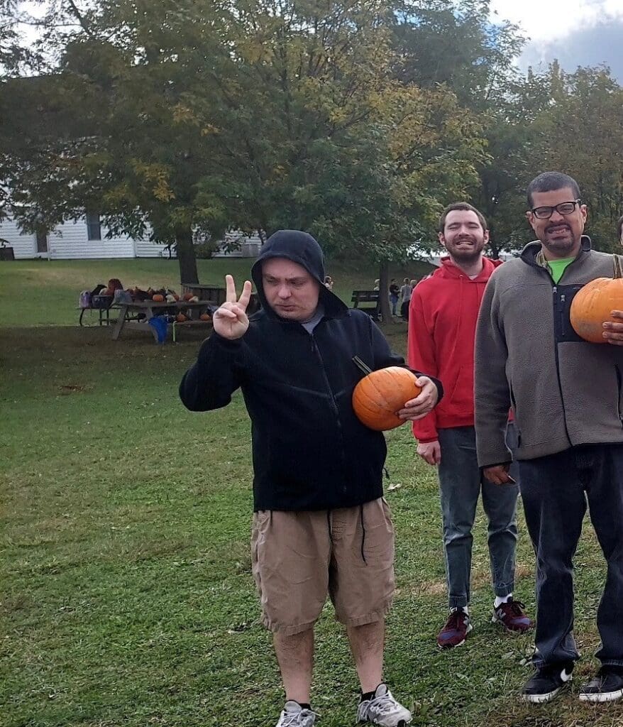 Three people outdoors holding pumpkins in a grassy area with trees.