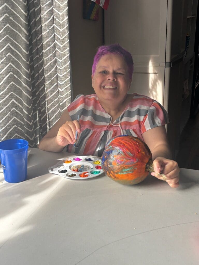A smiling woman painting a colorful pumpkin indoors.