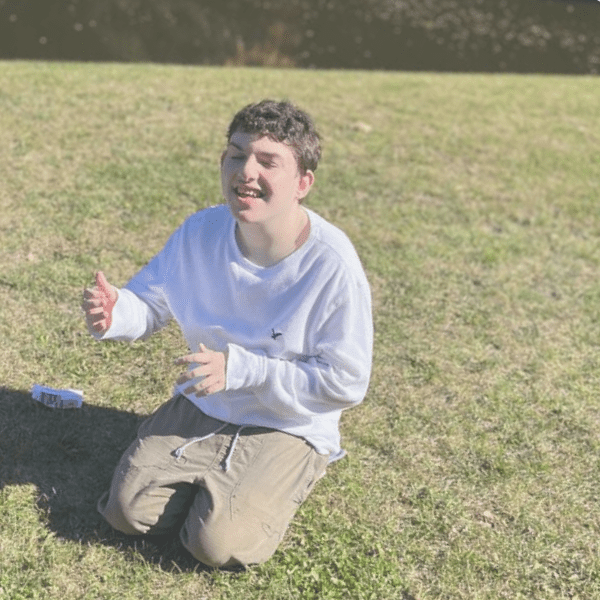 Person kneeling on grass, smiling and gesturing with hands.