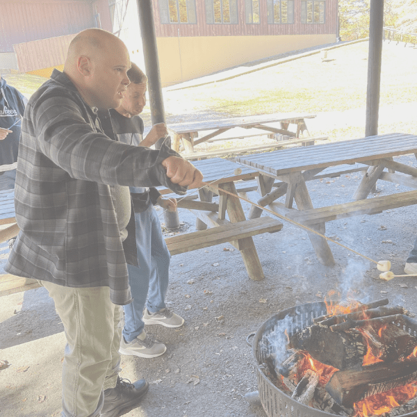 Two men roasting food over an open fire under a shelter.