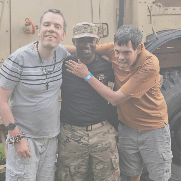 Three men smiling and embracing in a garage or workshop setting.