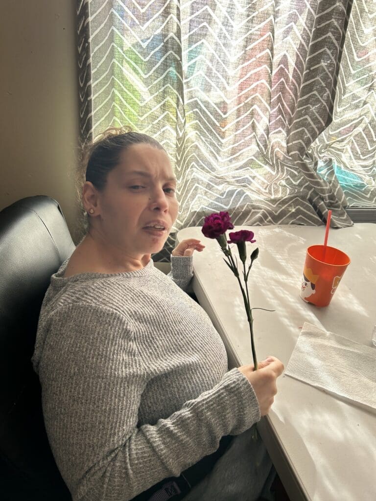 Woman holding two flowers while sitting at a table near a window.