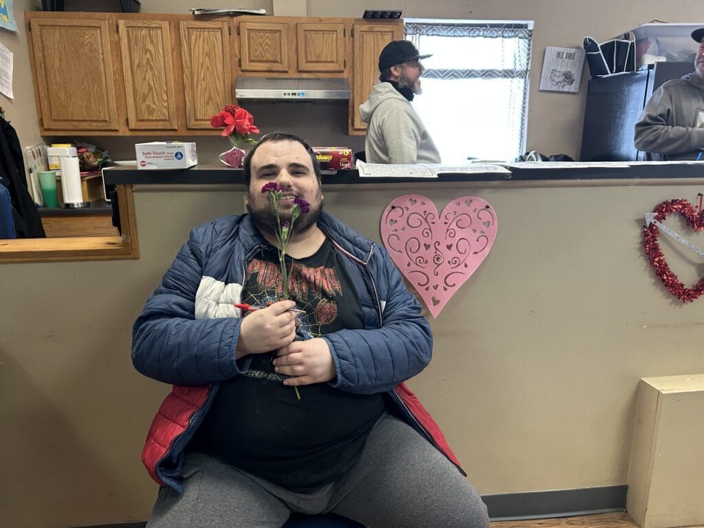 Man sitting indoors holding a small dog, with another person in the background.