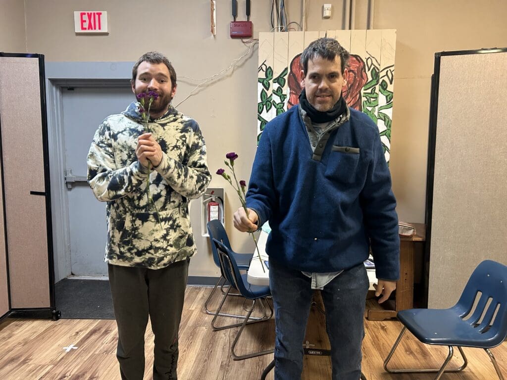 Two men standing indoors, one wearing a camouflage jacket and the other in a blue jacket.
