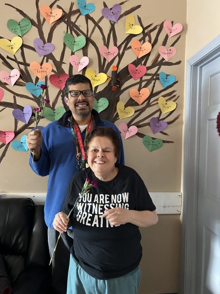 Two people smiling in front of a colorful handprint tree mural.