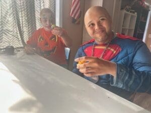 Two men dressed in superhero and pumpkin costumes smiling at a table.