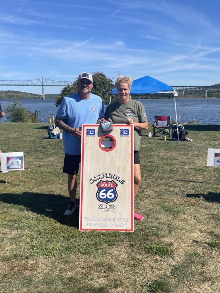 Two men posing with a vintage Route 66 cornhole board outdoors.