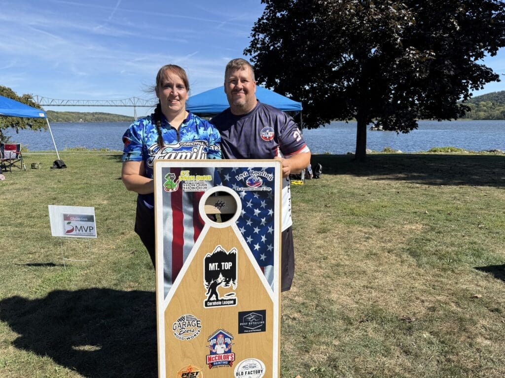 Two people standing outdoors by a cornhole board with a lake in the background.