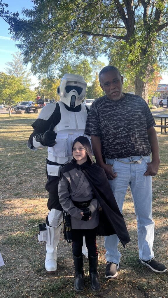 Three people posing outdoors, one in a stormtrooper costume giving thumbs up.
