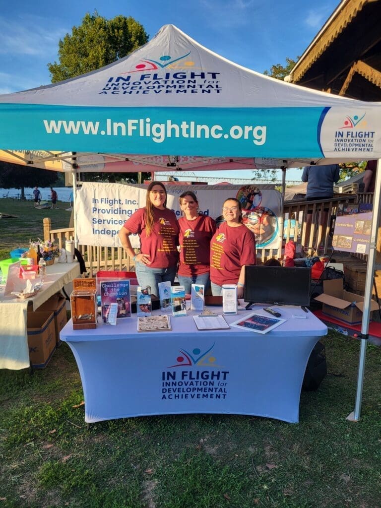 Three people at an In Flight Inc. booth with a blue canopy and informational materials.