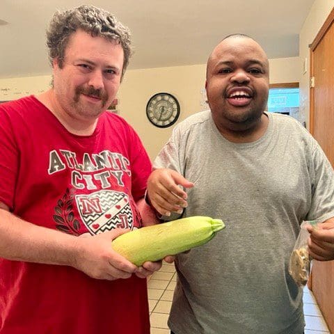Two men smiling, holding a large zucchini in a kitchen.