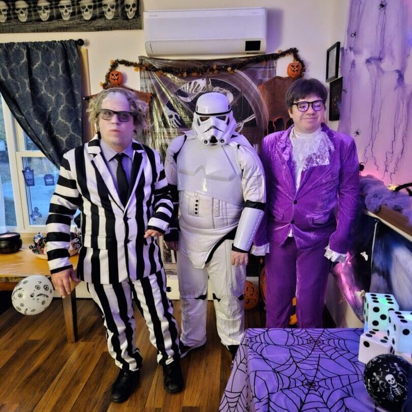 Three people in Halloween costumes standing indoors with decorations.